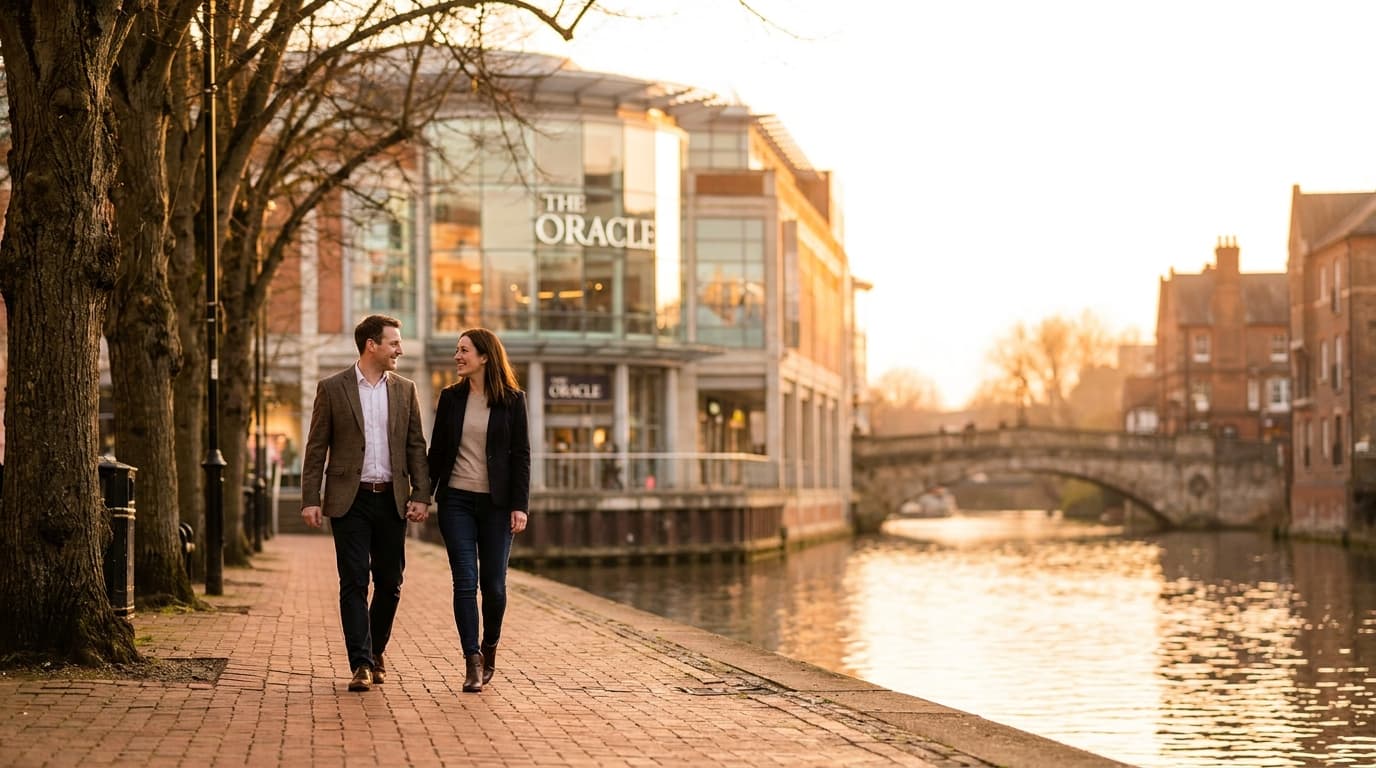 A couple walking hand-in-hand along the River Kennet at golden hour, with the glass and steel façade of the Oracle shopping centre reflected in the water behind them