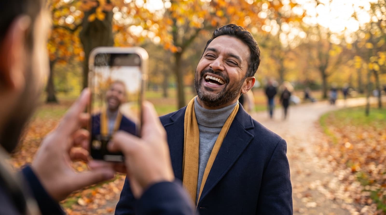 A laughing man with a short beard, wearing a navy coat and mustard scarf, has his photo taken on a smartphone by a friend in a park full of golden autumn leaves